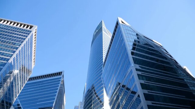 Modern glass office towers rise into clear blue sky in low-angle urban view, with reflective facades and angular architecture filling frame as camera subtly shifts upward