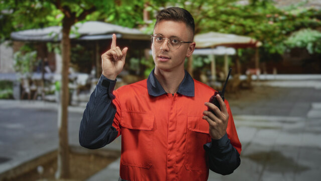 Young blond man in orange coverall holding a walkietalkie and points finger up on a street cafe terrace while wearing glasses and looking focused; alert readiness.
