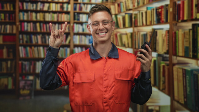 Man mechanic in orange coverall holding a walkie talkie and making rock horns gesture with his hand in a building library; playful joy.
