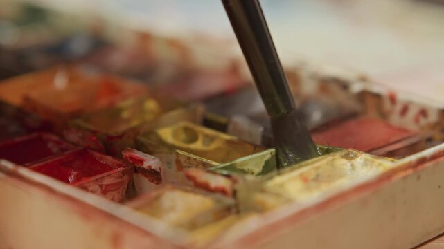 Close up of paintbrush mixing colors in watercolor palette with various hues. brush repeatedly dips into different compartments blending pigments while background remains softly blurred