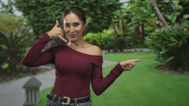 Woman wearing maroon off shoulder top showing call me hand gesture with left hand and pointing finger right with other hand in street pathway; playful invitation.