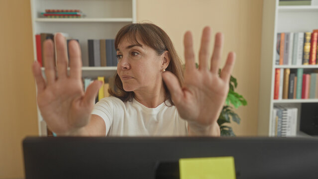 Woman holds palms forward above laptop at desk inside building, palms facing camera stop gesture; assertive boundary.