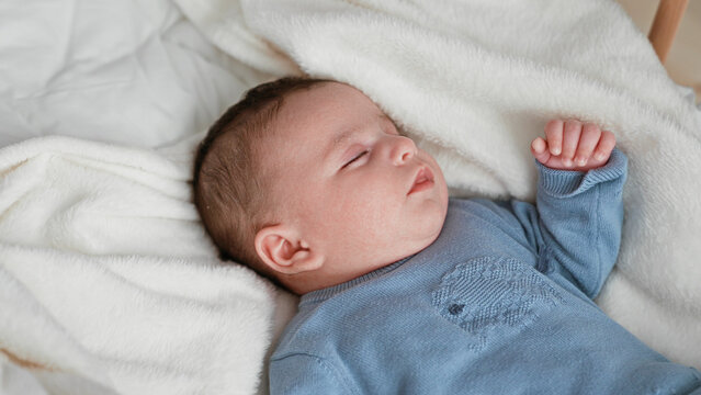 Newborn boy sleeping in a blue sweater with raised hand on a soft white blanket in a nursery cradle, eyes closed and relaxed; quiet serenity.