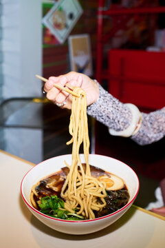 Vertical image of Asian woman hand holding chopsticks lifting noodles from ramen bowl in cafe. Visual ideal for Asian food marketing and restaurant promotion