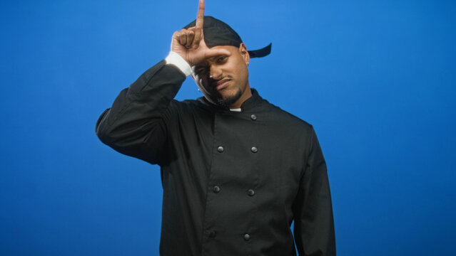 Man chef in black uniform points spatula at camera while making l sign on forehead in studio with blue backdrop; playful confidence.