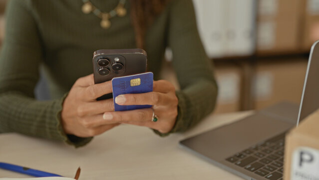Woman holding smartphone and creditcard in hands at a studio desk with parcels, laptop and notebook, tapping to process orders; small business focused.