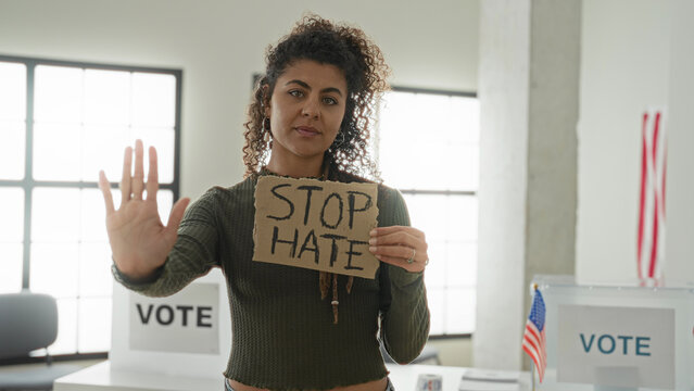 Woman holding cardboard sign reading stop hate while showing palm out toward camera inside a voting building with vote booths and us flags visible; solidarity protest.