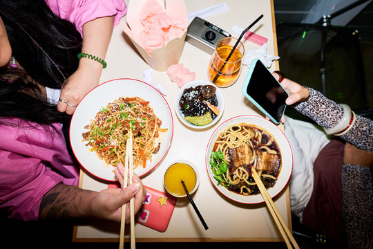 Top view shot of young adult Asian friends eating Asian food in cafe and using smartphone. Image represents modern youth lifestyle, suitable for food delivery app marketing