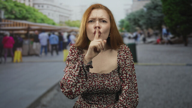 Redhead woman pressing index finger to lips for silence while standing in a crowded urban street market with blurred pedestrians behind her; warning secrecy.