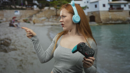 Young woman wearing cat ear headphones holds gamepad and points finger in front of seaside building while pouting and squinting in displeasure  frustration defiance. © Krakenimages.com