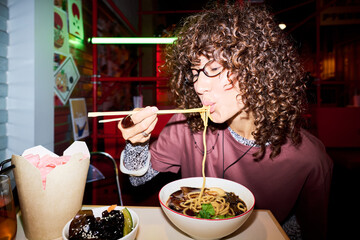 Young adult Asian woman eating ramen noodles with chopsticks in Asian restaurant. Image represents...