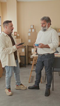 Man holding tablet gestures with hand to colleague holding coffee amid stacked parcels in building; collaboration.