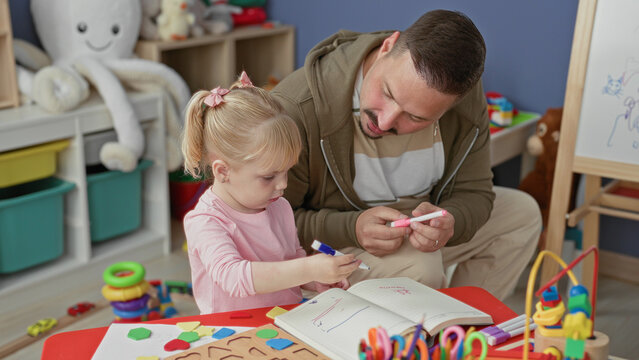 Man guiding a girl holding markers and drawing in a notebook at a colorful play table with toys in building; learning warmth.