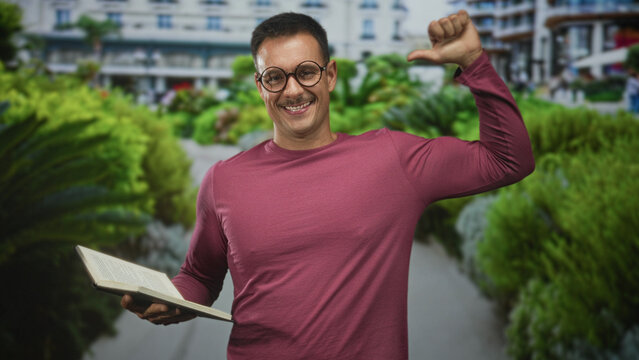 Young hispanic man with moustache and glasses holding an open book and pointing thumb to chest while smiling in front of building; confidence curiosity.