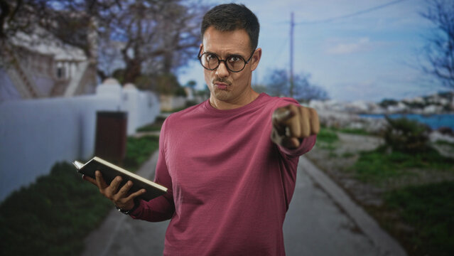 Hispanic man with moustache and glasses holds book and beckons with hand on a street by the sea; thoughtful invitation.
