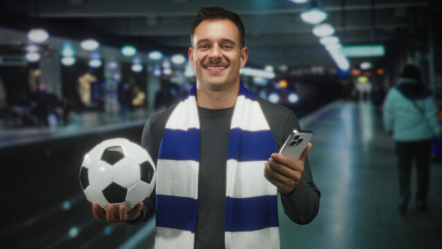Man with moustache holding soccer ball and smartphone, palm presenting ball, wearing striped scarf in subway station; matchday excitement.