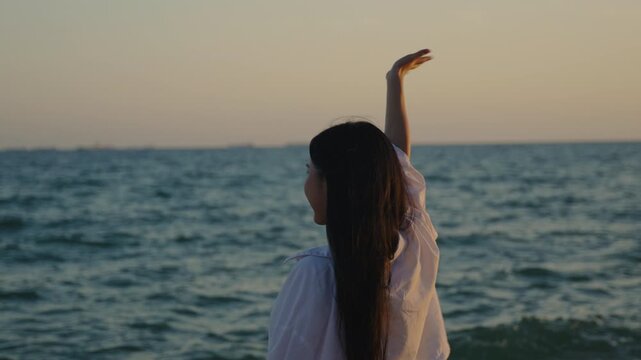 Back view of Asian woman on beach waving hand in goodbye gesture to ocean, looking at calm sea water with arm raised, enjoying peaceful travel vacation during beautiful sunset.