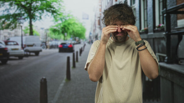 Man covering his eyes with hands on a city street, wristwatch and bracelets visible, rubbing face gesture; fatigue commute routine.