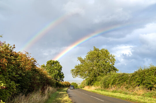 Car driving along a rural road beneath a bright double rainbow