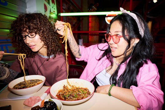 Young Asian women eating noodles in Asian restaurant, sharing food and smiling. Image represents youth lifestyle and is suitable for food delivery app marketing, medium shot