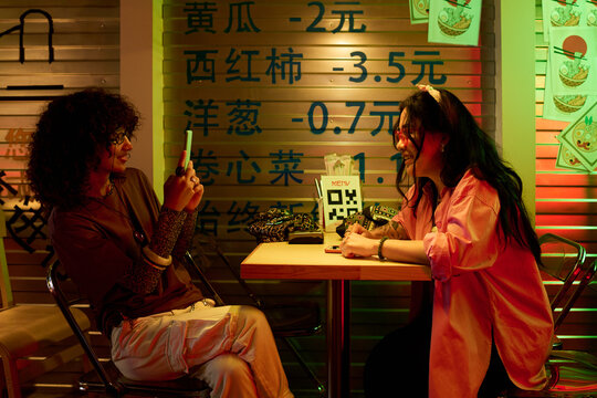Young adult Asian women sitting at cafe table, one taking photo of other with smartphone. Image represents youth lifestyle and is suitable for social media marketing campaigns