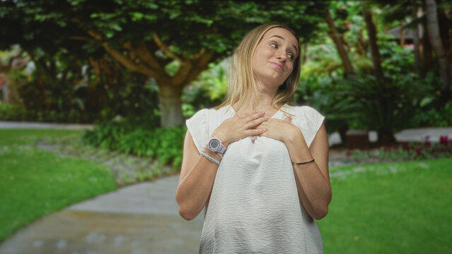 Woman points thumb to herself in white blouse with wristwatch on park path in forest, playful smirk; self confidence pride.