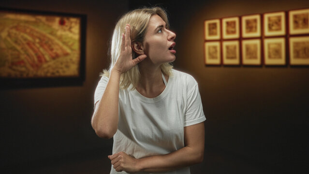 Woman, young blonde, with crossed arms and gesturing hand toward framed art in gallery; quiet contemplation.