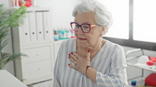 Elderly woman holding chest with hand in clinic building wearing glasses and striped blouse; concern vulnerability.