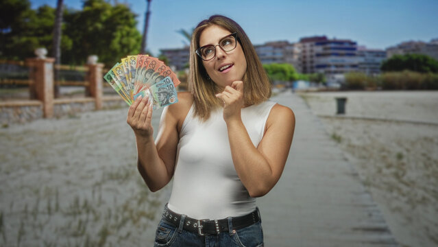 Woman holding a fan of australian dollar banknotes and touching chin on a street; thoughtful money curiosity.