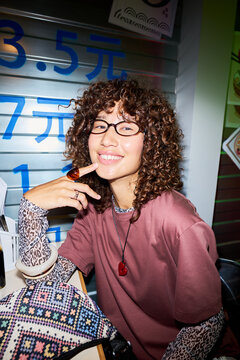 Vertical portrait of smiling young Asian woman with curly hair sitting in cafe. Image is suitable for youth lifestyle marketing, food blogging, and social media campaigns