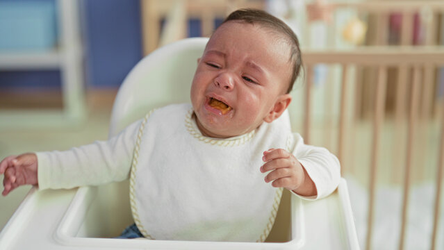 Baby boy crying and spitting spoonful of puree into mouth with messy bib while seated in highchair in building; feeding frustration.