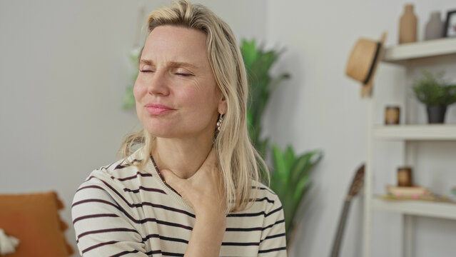 Woman rubbing neck with hand in a living room, hand pressed to side of neck while sitting; relief calm recovery.