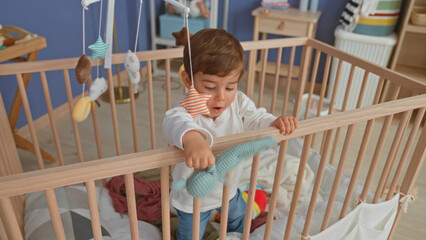 Boy stands in wooden crib and reaches for hanging plush mobile while gripping the rail inside a domestic building  gentle curiosity. © Krakenimages.com