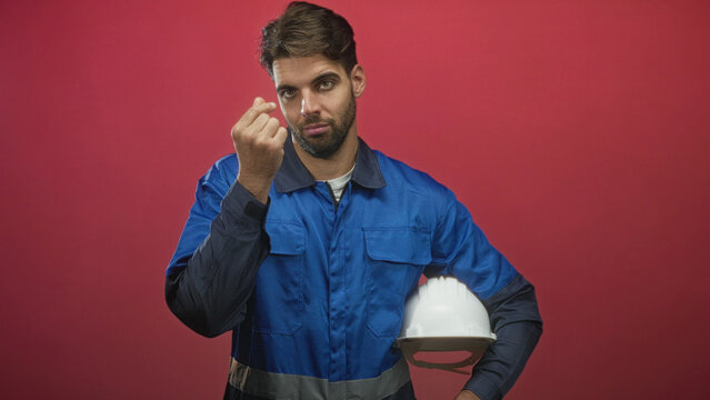 Hispanic bearded man in blue work overalls holds white hardhat and makes pinch fingers money gesture in studio; confidence professionalism.