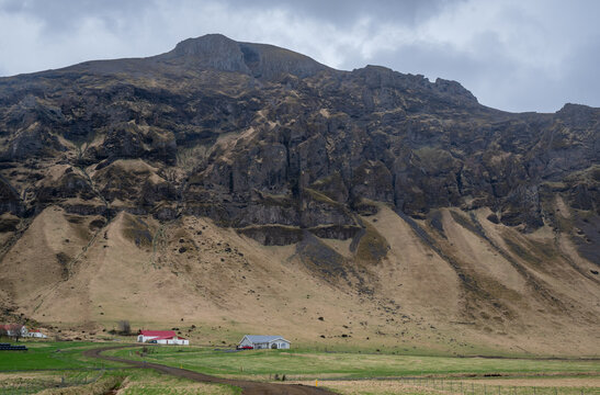 Rugged mountain landscape in Iceland rural houses situated at the base of steep hills. Low clouds over peak of mountain. Farmland Icelandic landscape.