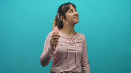 Fototapeta premium Young woman holds pink soft serve ice cream cone in hand, looking up and smiling against a turquoise studio wall; playful joy.