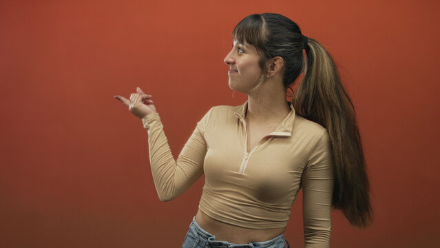 Young woman smiling and flicking her finger with pointing hand, visible midriff against orange studio backdrop; spontaneous joy.