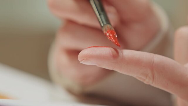 Close up of person painting their finger red. hand holds paintbrush applying bright orange red pigment to extended index finger with steady brushstrokes. Soft focus background emphasizes