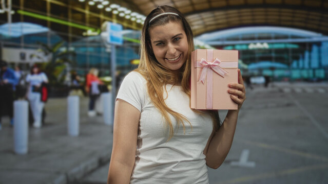 Woman holds pink present against cheek while smiling and tilting head at airport; happy celebration.