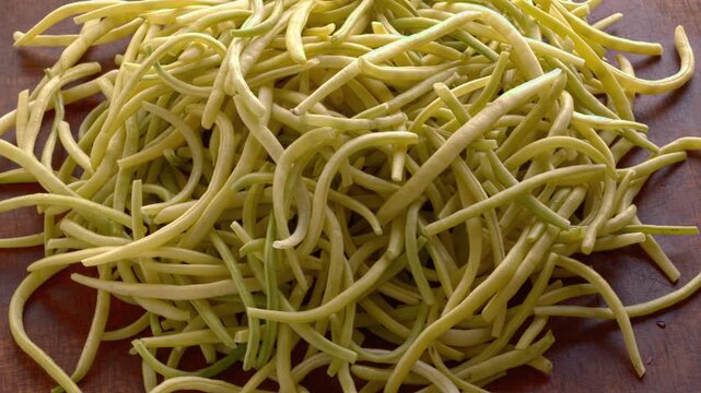 A heap of Golden Teepee yellow wax beans rotating on brown plywood. Fresh, organic, raw, trimmed bean pods pile close-up on a dark wood background, slowly spinning in a tilted view.