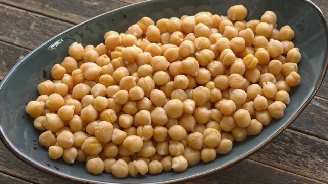 Boiled chickpeas heap rotating on an oval ceramic platter. Fresh organic garbanzo beans, close-up on a rustic dark wood plank background, slowly spinning in a tilted view with natural window light.