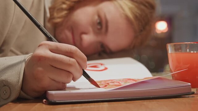Close up of young woman painting with watercolors in art journal focusing on brush strokes and vibrant colors. glass of pink drink sits nearby as woman works intently at table capturing