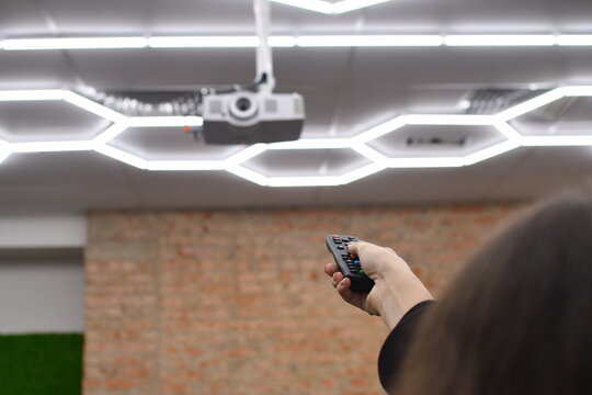 Back view woamn using remote control to operate ceiling projector and modern LED lighting system in conference room. Selective focus.