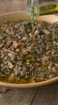 Pouring an olive oil drizzle from a bottle onto a thick, homemade, traditional Mediterranean soup. Greek Faki me Seskoula lentil stew with Swiss chard on rustic wood. Vertical video.