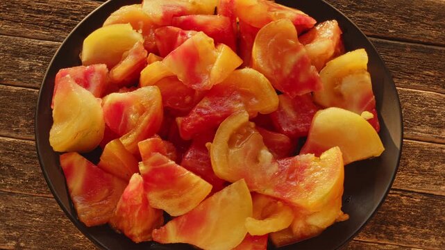 Old German tomato wedges rotating on a black plate. Sliced organic orange-red heirloom tomato chunks on a rustic dark wooden background in warm late-afternoon golden light.