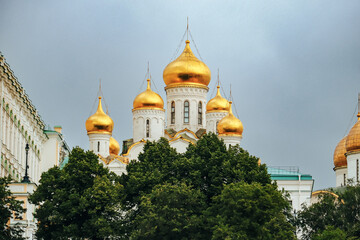 Golden onion domes of Orthodox cathedral rising above green trees under overcast sky in Moscow Kremlin, with cathedral © Val MAK