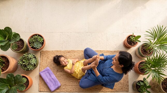 Top view of a happy Indian mother playing with and massaging her toddler daughter on a jute rug surrounded by potted plants at home