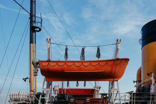 An orange lifeboat hanging on davits on a ship deck near a large yellow funnel under a blue sky with soft clouds.