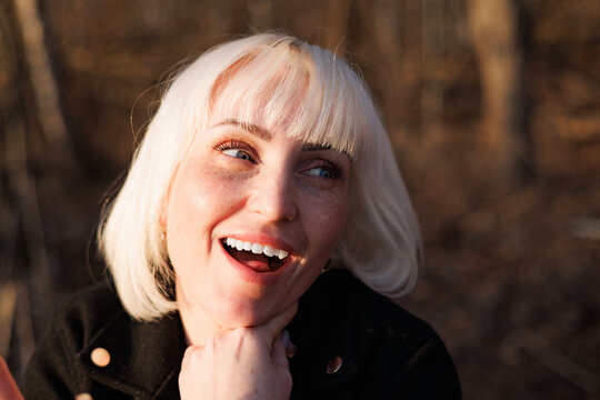 A joyful smiling young woman with blonde hair looking away in a forest during a warm golden hour sunset in autumn.
