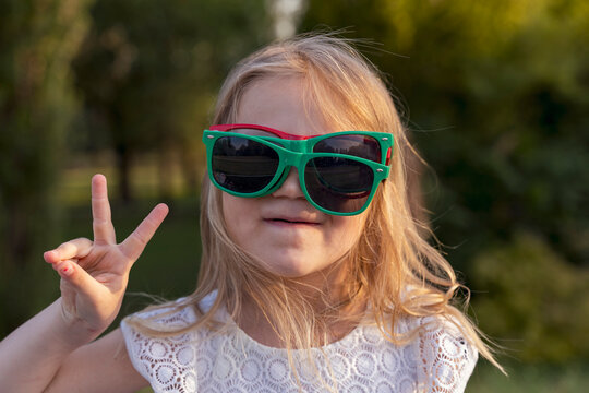 Young blonde girl wearing two pairs of sunglasses and showing peace sign outdoors